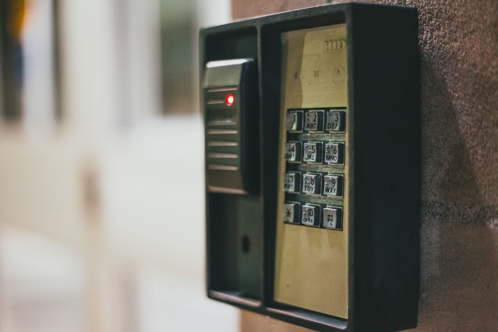 A black wall-mounted access-control unit — a numeric keypad with keys labelled with letters beside a card-reader panel with a small red indicator light — fixed to a textured brown wall with a blurred corridor behind, indoors in low light.