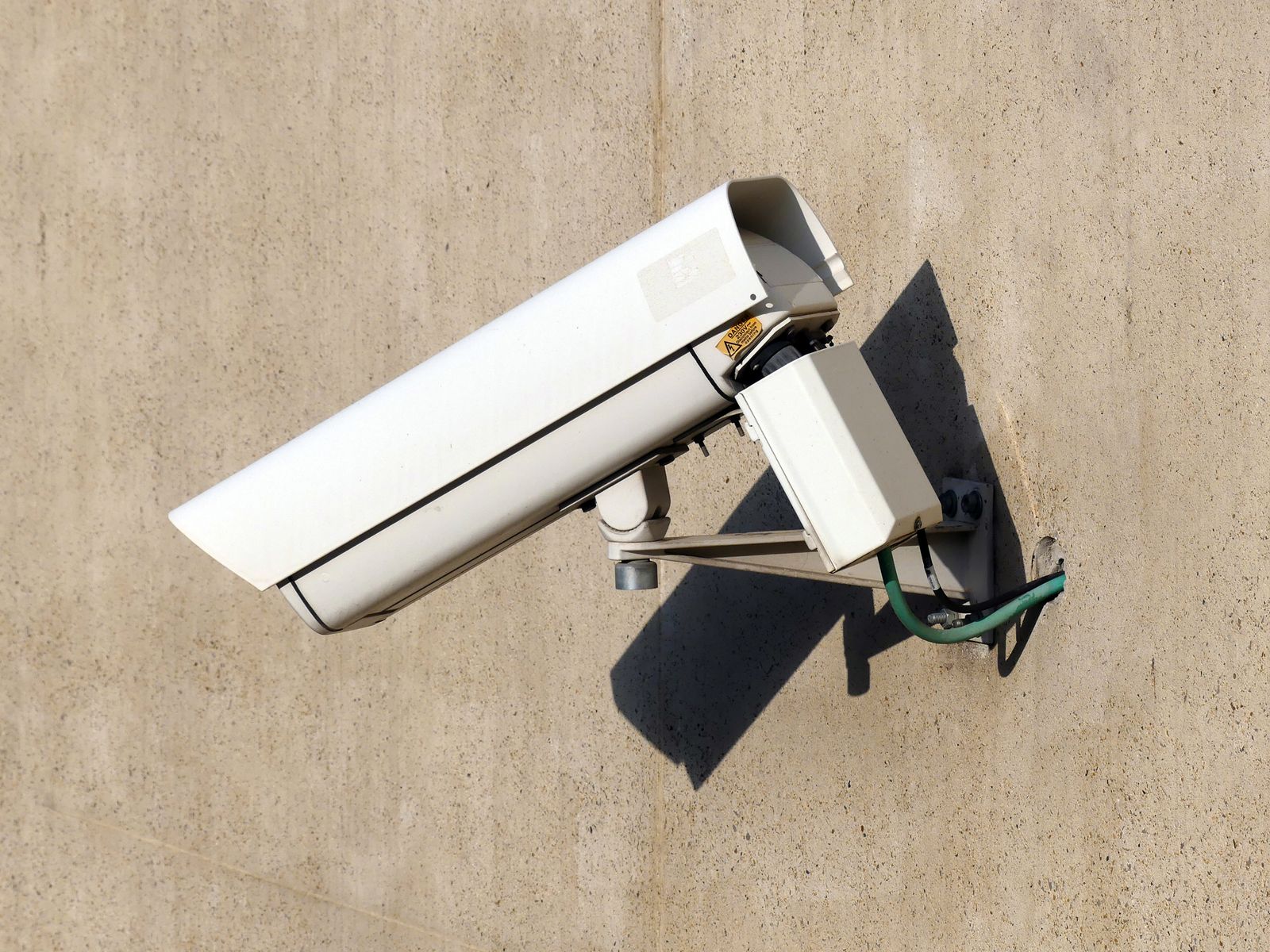 A white box-style CCTV camera in a weather housing with a sun shield, mounted on a bracket against a plain concrete wall, angled down and to the right and casting a hard shadow in bright daylight, with a small yellow warning sticker on the housing.