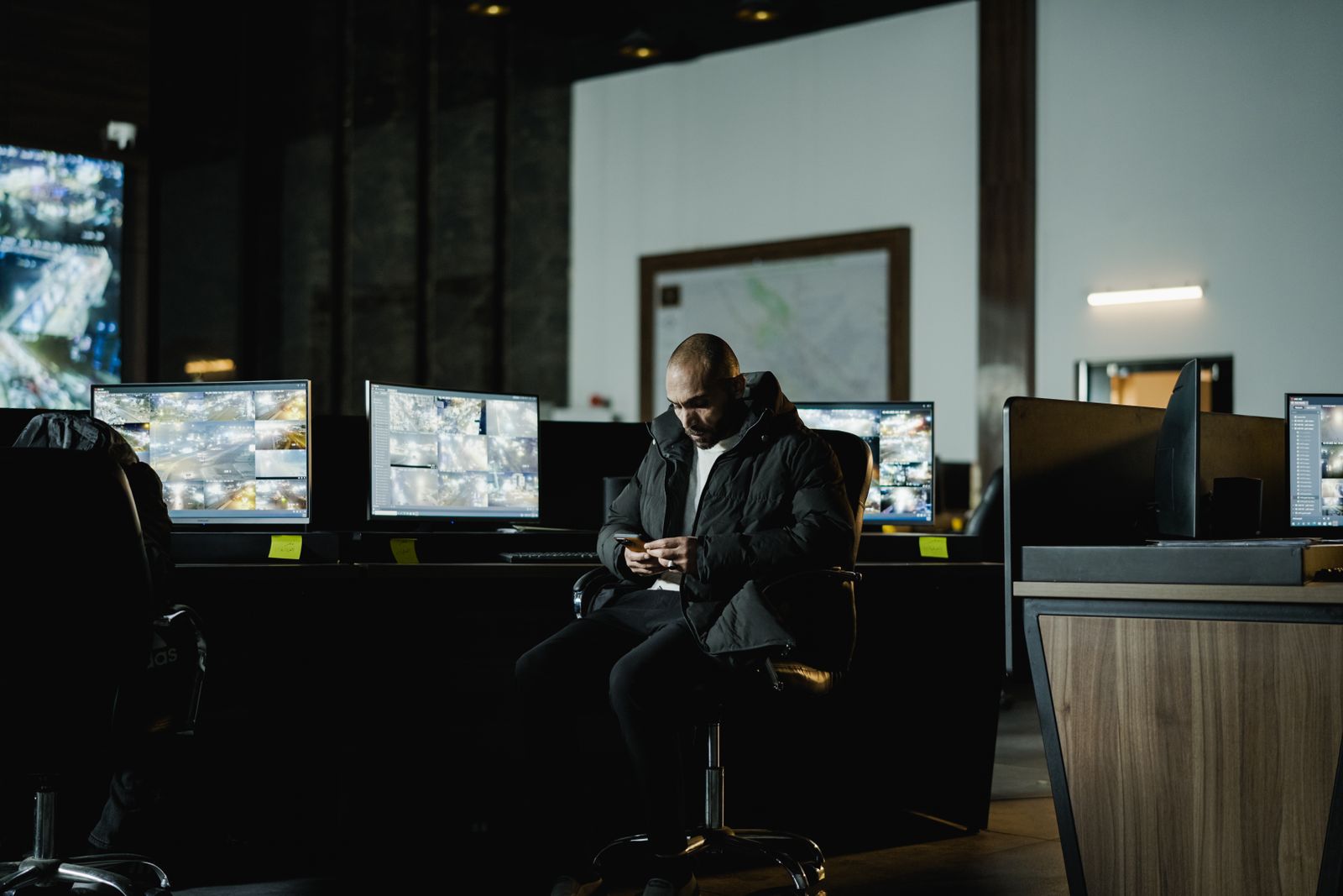 A dimly lit monitoring room where a man in a dark padded jacket sits at a long desk holding a phone, with several large monitors showing multi-camera CCTV grid views, a framed area map and a strip light on the wall behind, in low light.