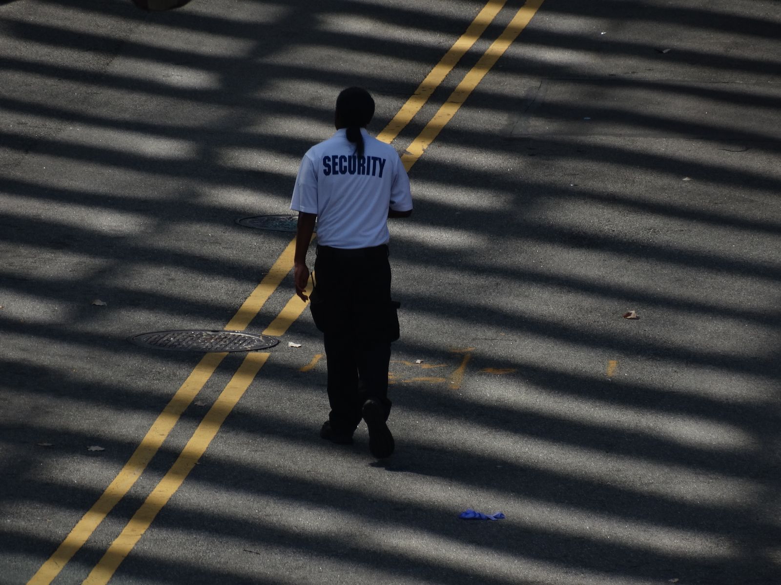 A lone security officer walking across a wide tarred road marked with a double yellow line, seen from behind and above with long tree shadows striped across the road; the officer wears a pale short-sleeve shirt printed "SECURITY" across the back and dark cargo trousers, in daytime.