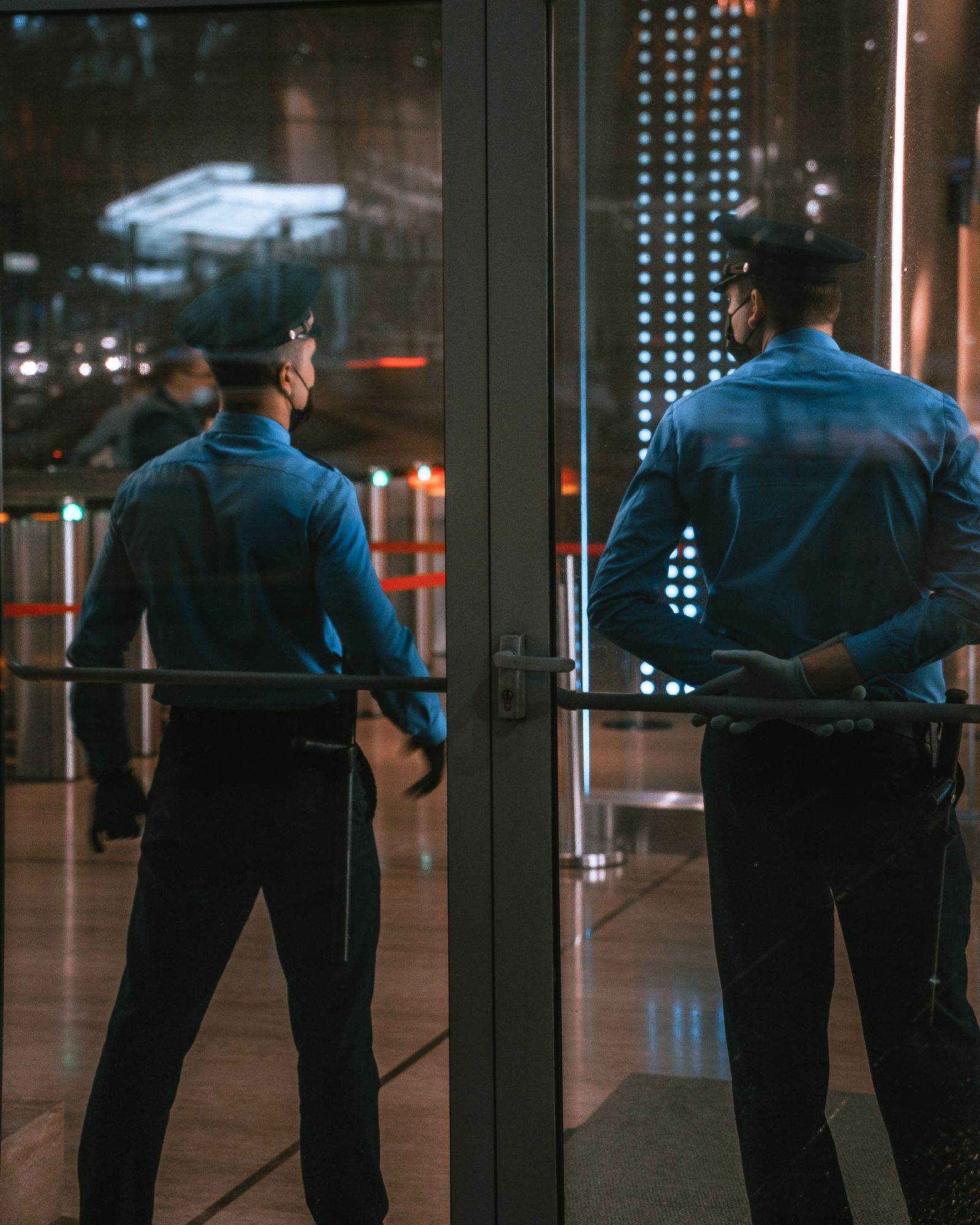 Two uniformed security officers seen from behind, standing at a glass entrance door with metal turnstiles and a roped queue lane behind them, in blue button-up shirts and peaked caps under evening interior lighting.
